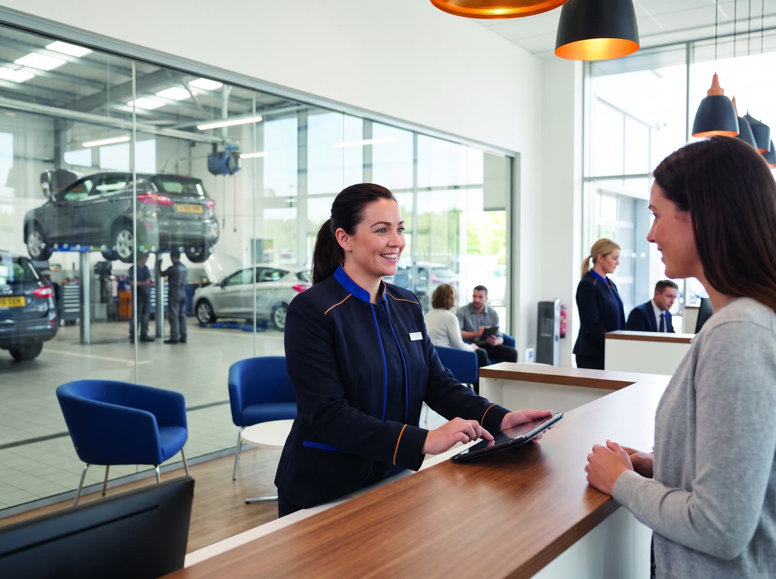 A service advisor at a dealership service desk taking a booking next to the workshop
